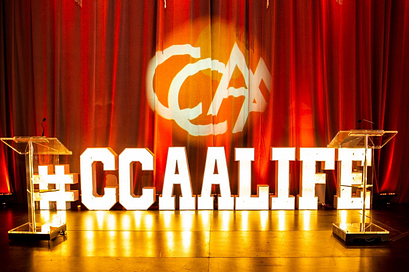 a stage with large letters that say CCAA life and the CCAA logo projected on the curtain on the back wall