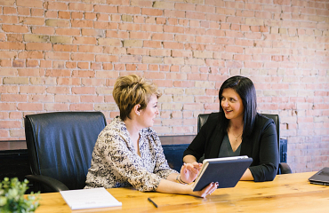 two women smiling in a boardroom while looking at a book