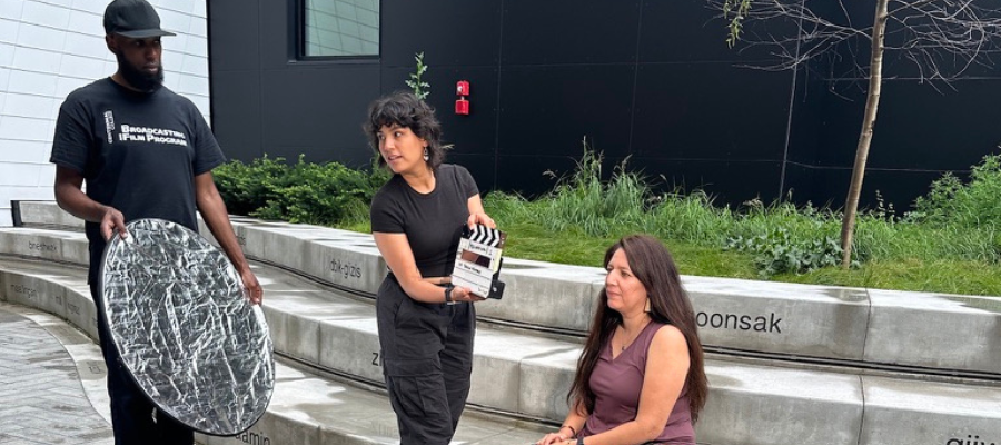 two film crew members, one holding a reflector and one holding a clapperboard, prepare to film a seated person