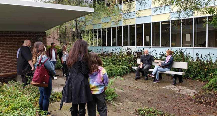Kit Hood sitting with Linda Schuyler on a Memorial Bench while multiple people observe them