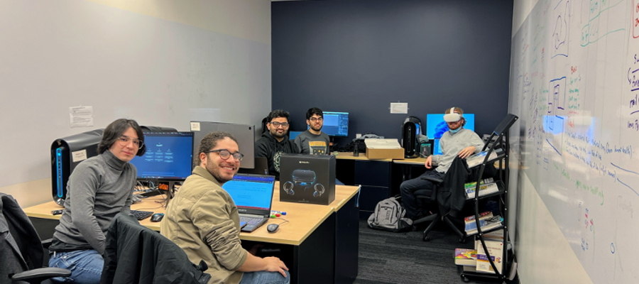 five centennial students in a computer room, one of the student is wearing a VR headset, whilst the rest look back at the camera smiling