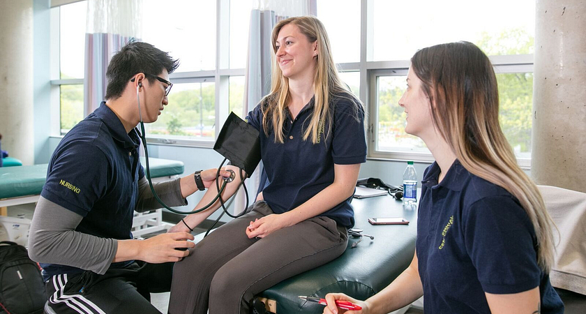 Picture of three nursing students in the classroom checking a woman's pulse with a stethoscope
