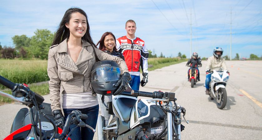Picture of three students standing in a line with two motorcycles in the forefront. Two students are riding in the background