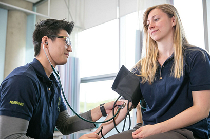 Nursing student wearing dark blue scrubs checking another student's blood pressure